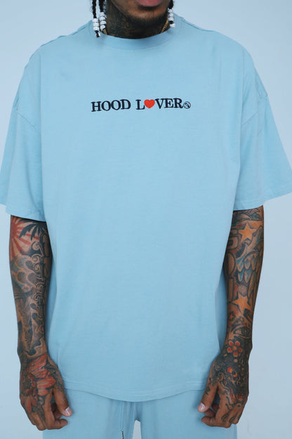 Oversized graphic streetwear t-shirt in light blue worn by a model with tattoos, featuring a stylish 'HOOD LOVER' graphic.