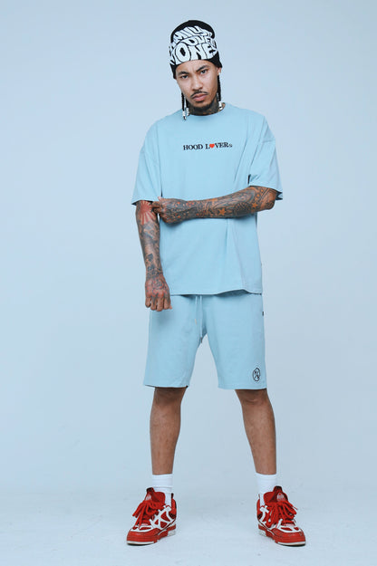 Model wearing an oversized graphic streetwear t-shirt with relaxed fit in light blue, paired with matching shorts.