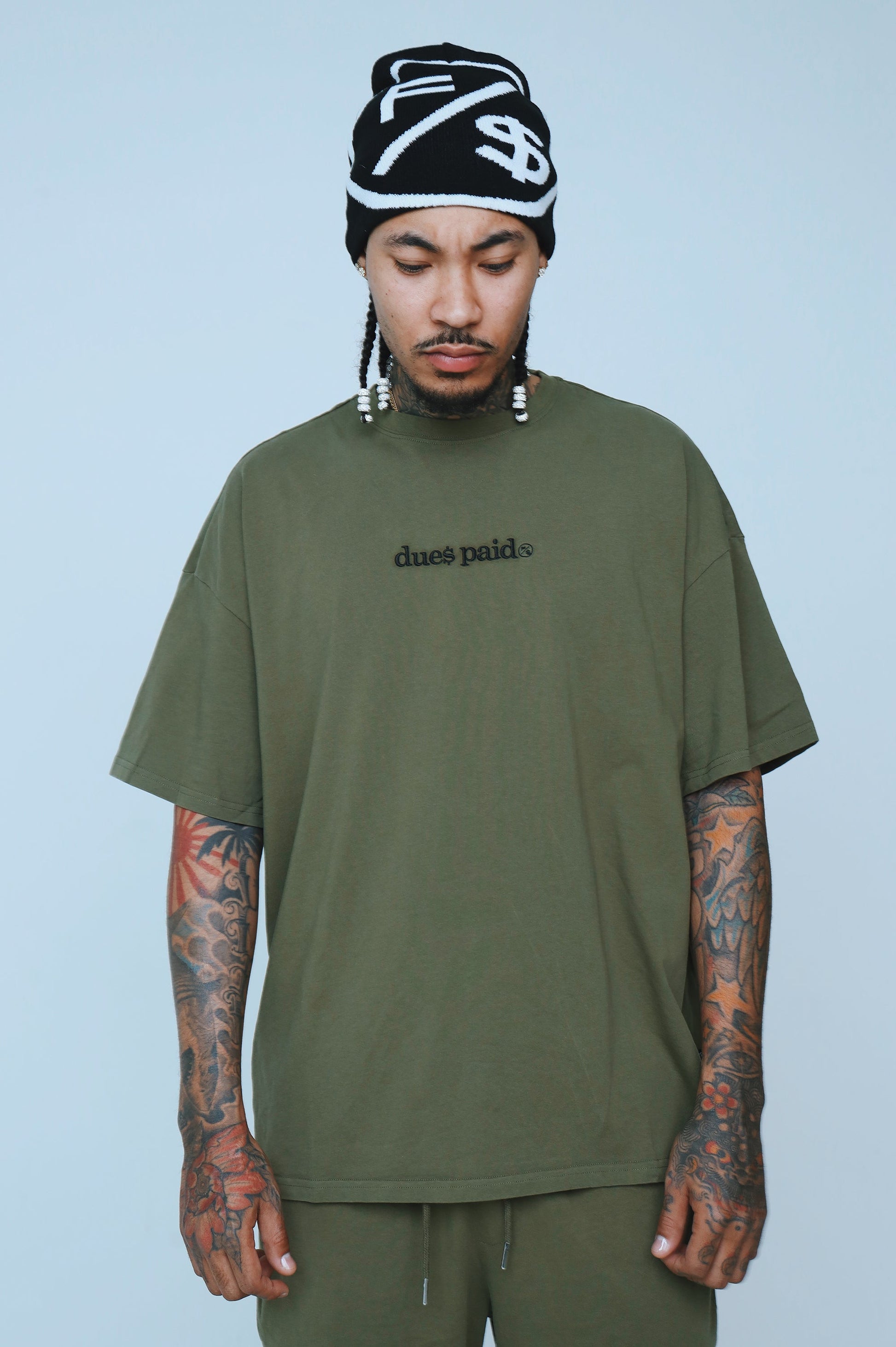 Model wearing oversized graphic t-shirt streetwear in olive green with 'dues paid' text, showcasing relaxed fit.