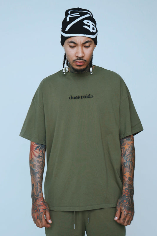 Model wearing oversized graphic t-shirt streetwear in olive green with 'dues paid' text, showcasing relaxed fit.