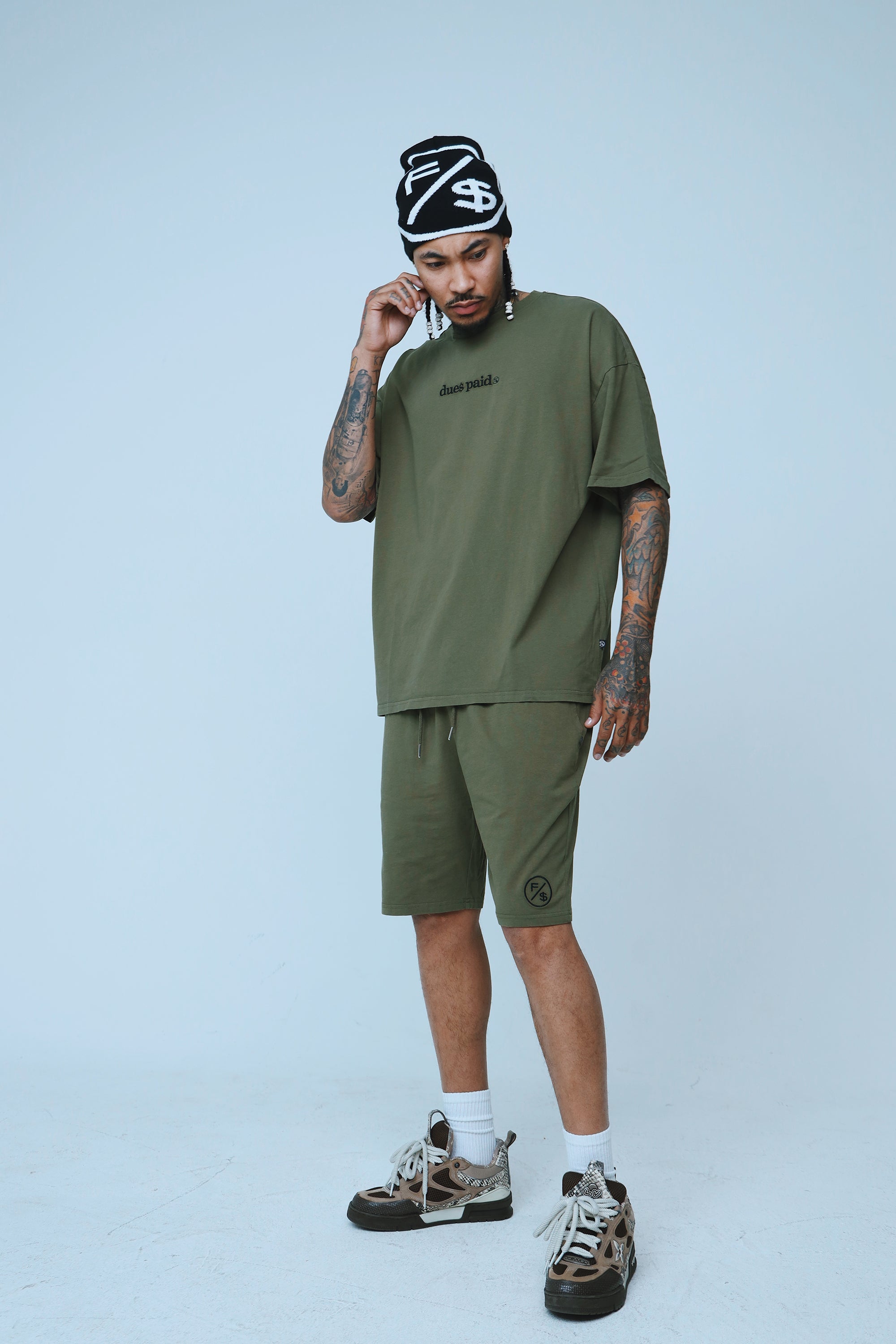 A model wearing oversized streetwear shorts in olive green with a relaxed fit, styled with a matching oversized tee and beanie.
