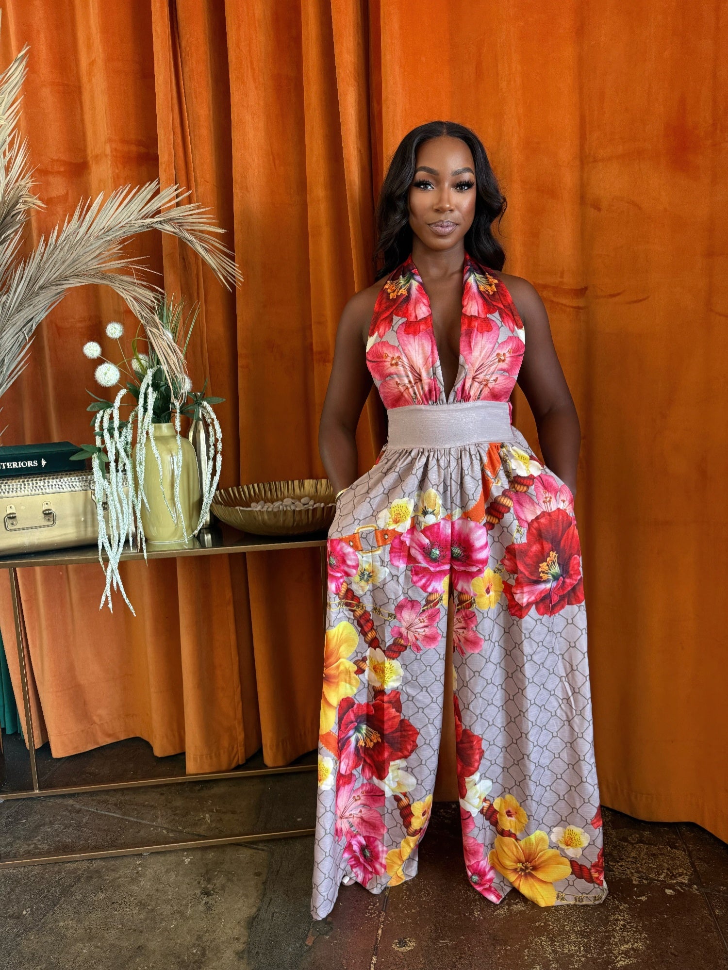 A model wearing a tropical floral jumpsuit with a plunging neckline and wide-leg silhouette, styled against an orange backdrop.