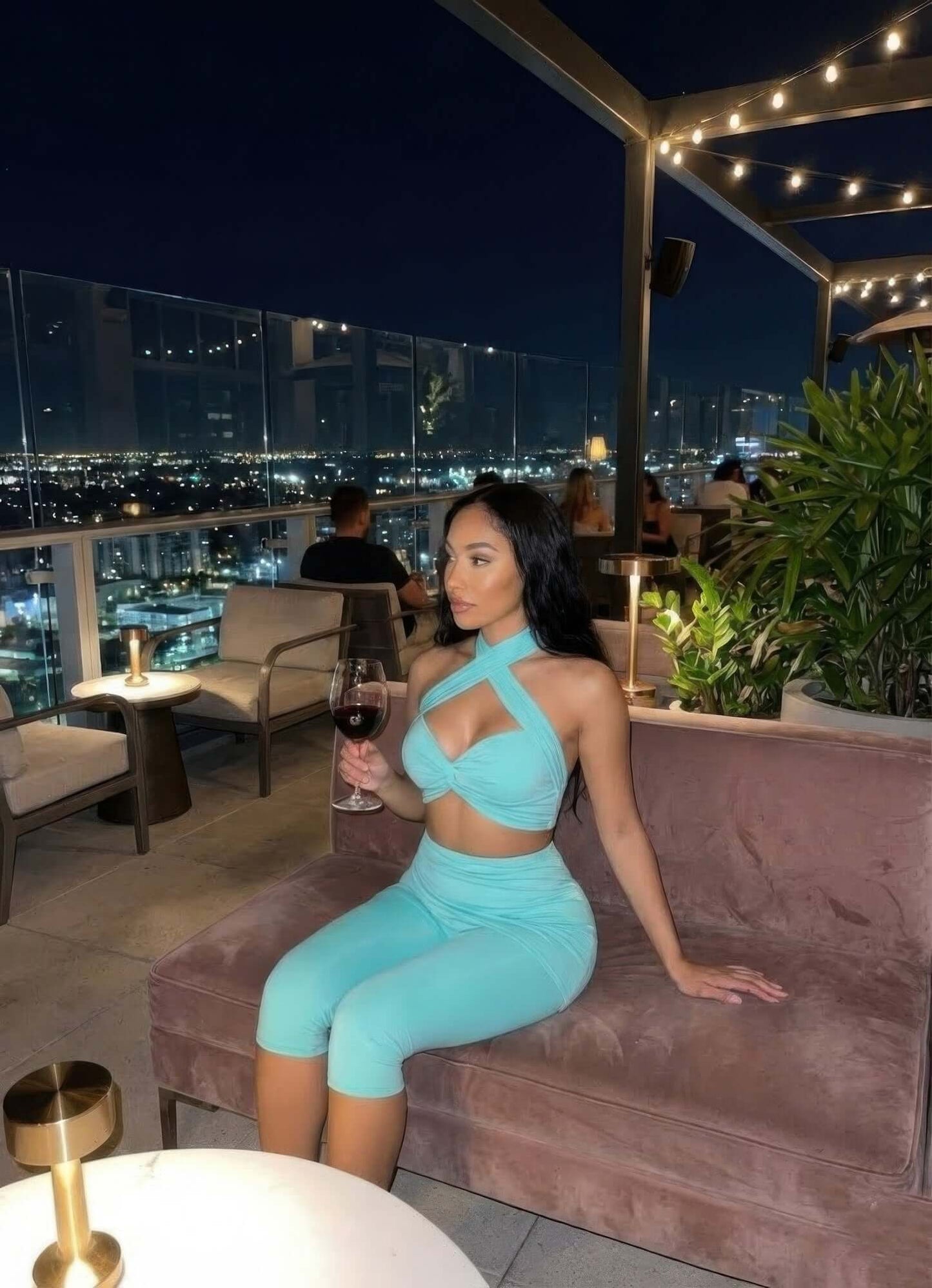 Woman wearing Crisscross Halter Two Piece Set in aqua with twist-knot front, seated on rooftop lounge at night holding wine