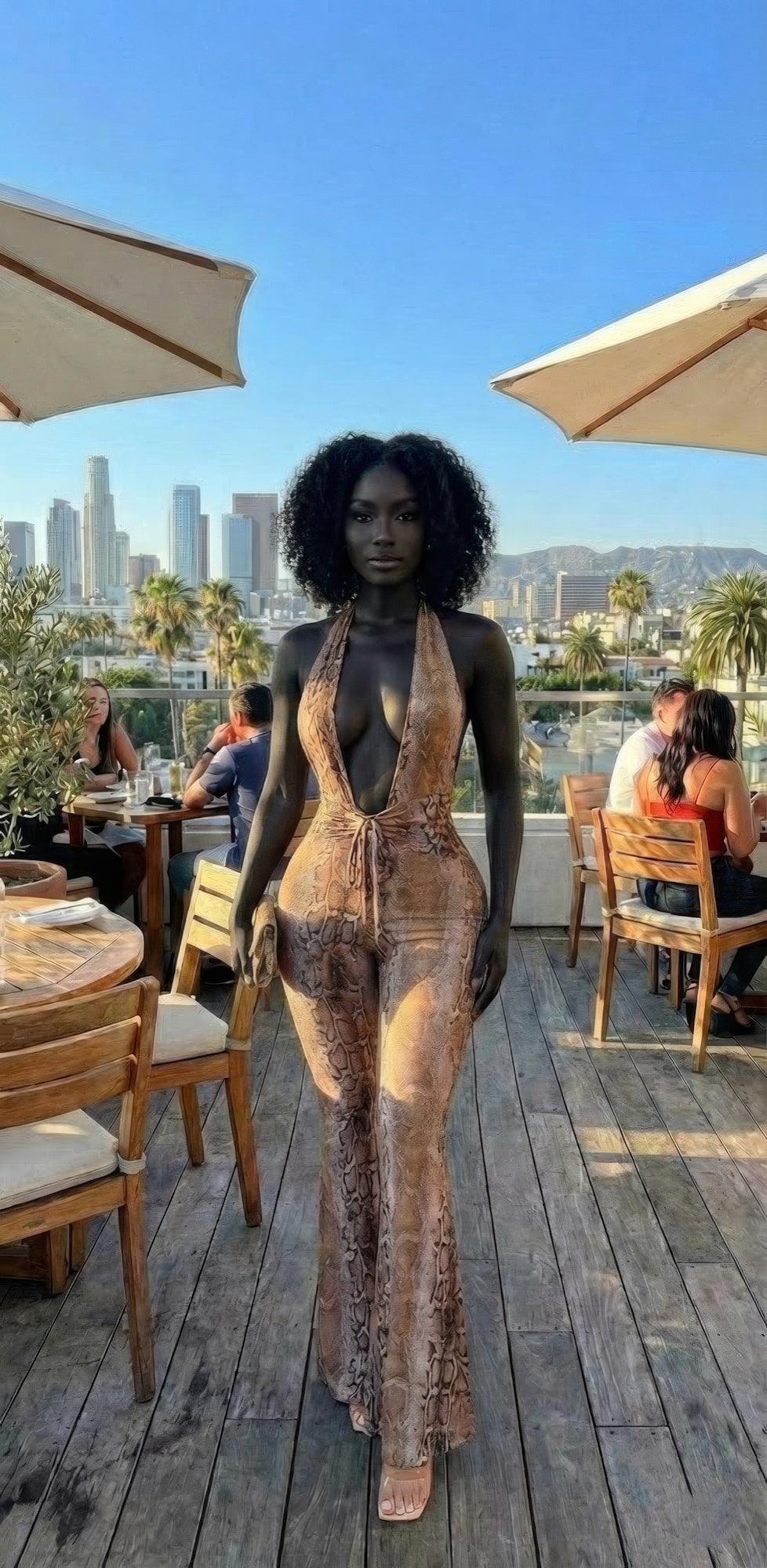Snake Print Tie Open Back Jumpsuit worn by woman, deep V halter and flared legs on outdoor patio with city skyline backdrop