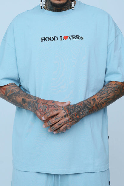 Oversized graphic streetwear t-shirt in blue worn by a person with tattoos, showcasing relaxed fit and urban style.