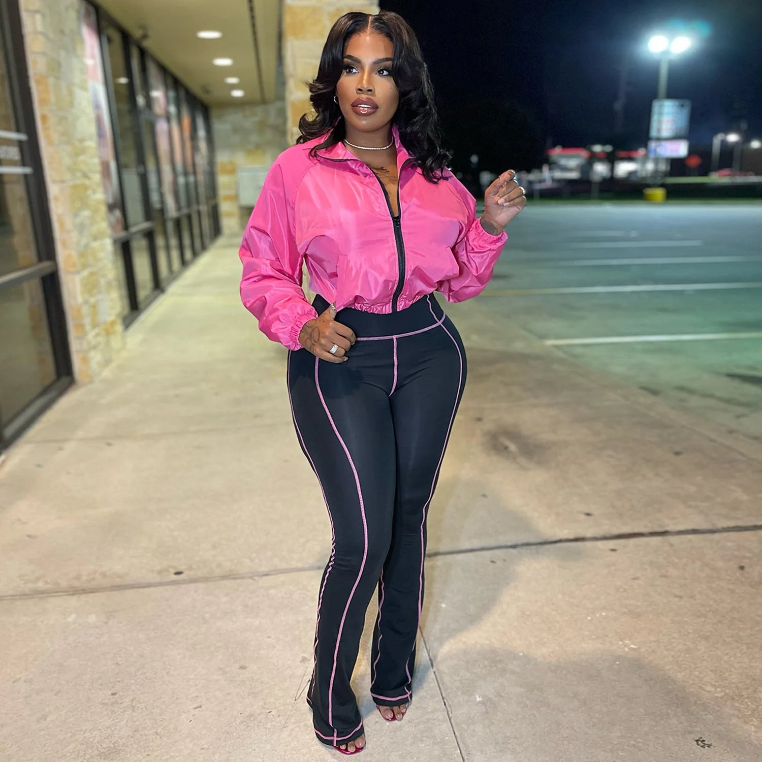 Woman wearing a glossy pink cropped jacket and black contrast stitch leggings in the Velocity Luxe Zip-Front Active Set.