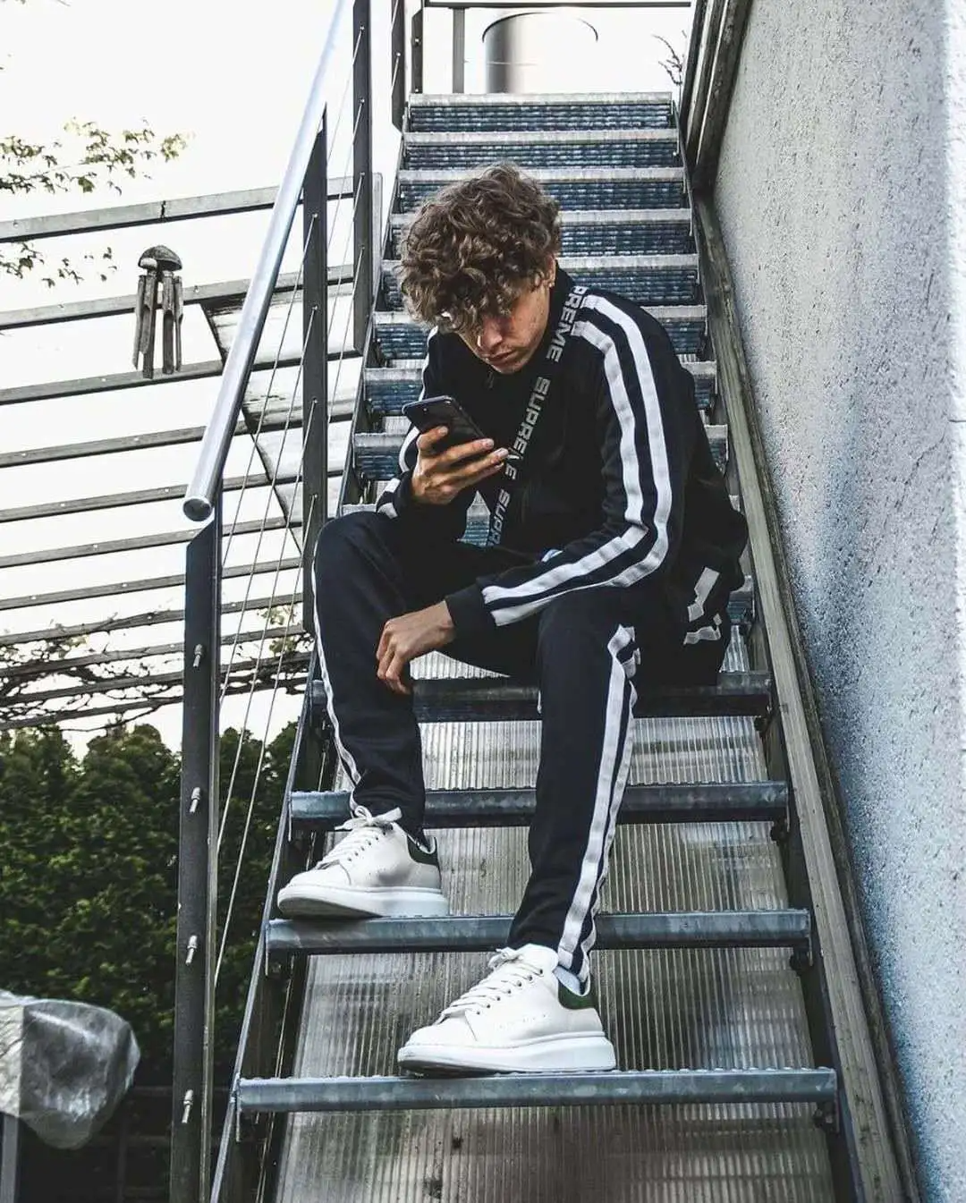 Young man in men's striped jogger set sitting on stairs, looking at phone in casual street style.