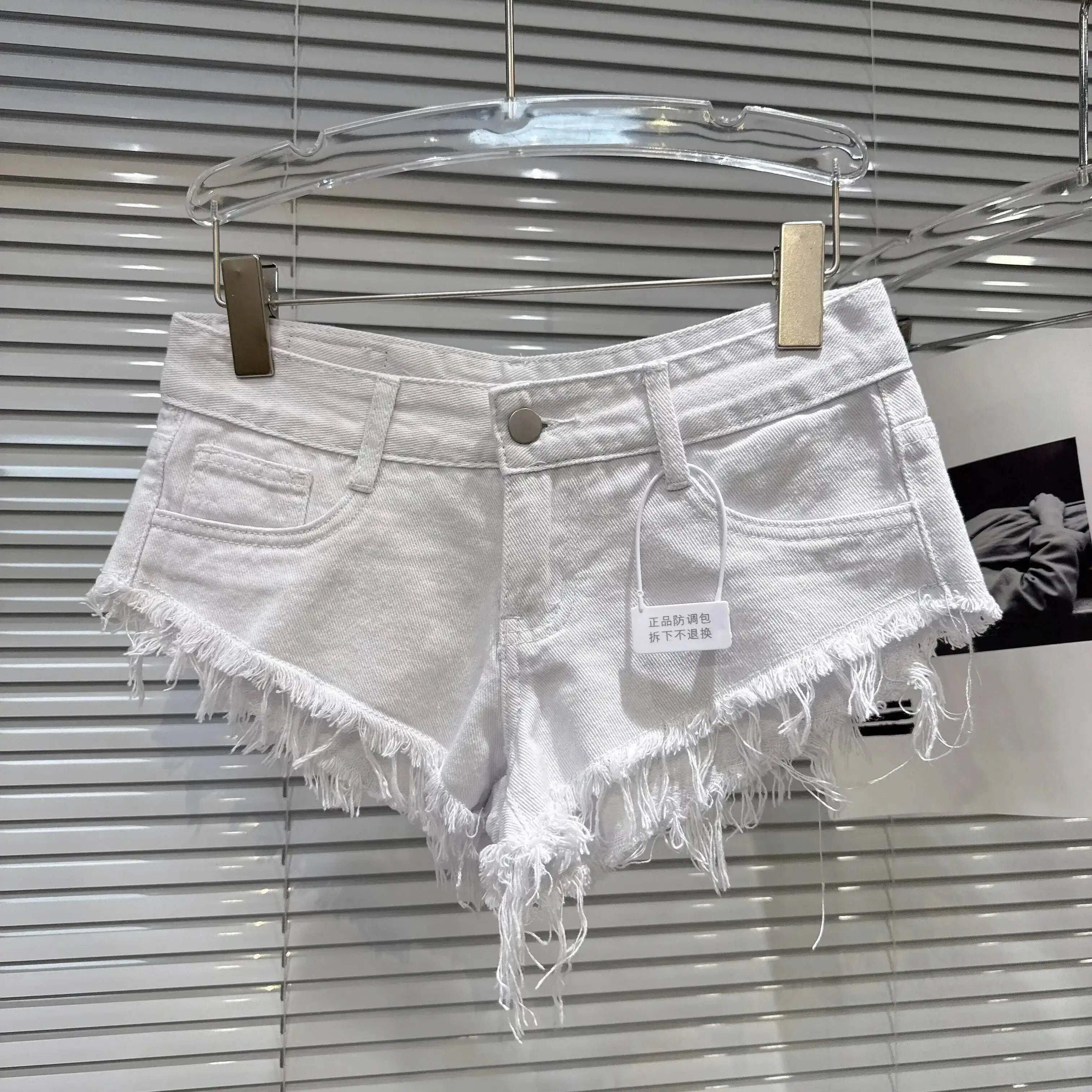 Distressed Raw-Hem Low-Rise Denim Shorts in white with frayed hem and low-rise waist hanging on display.
