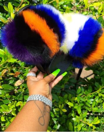 Colorful plush faux fur slides held in hand against a background of green foliage, showcasing vibrant purple, orange, and blue fur.
